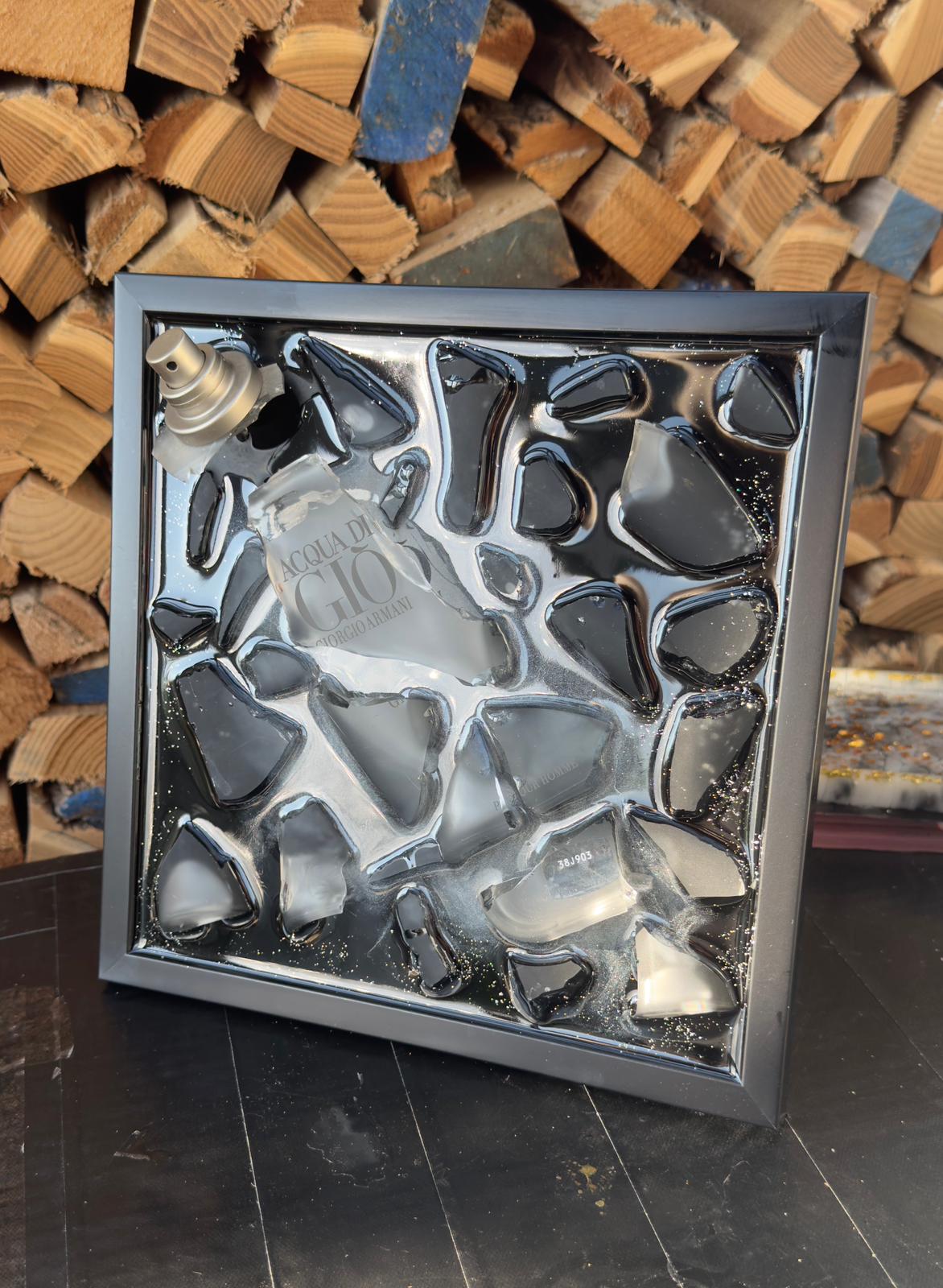 Gerahmtes modernes Kunstwerk aus zersplittertem schwarzen Parfumflakon, arrangiert als dreidimensionale Upcycling Wandkunst mit glänzenden Glasfragmenten in einem schwarzen Rahmen.