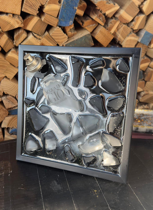 Gerahmtes modernes Kunstwerk aus zersplittertem schwarzen Parfumflakon, arrangiert als dreidimensionale Upcycling Wandkunst mit glänzenden Glasfragmenten in einem schwarzen Rahmen.