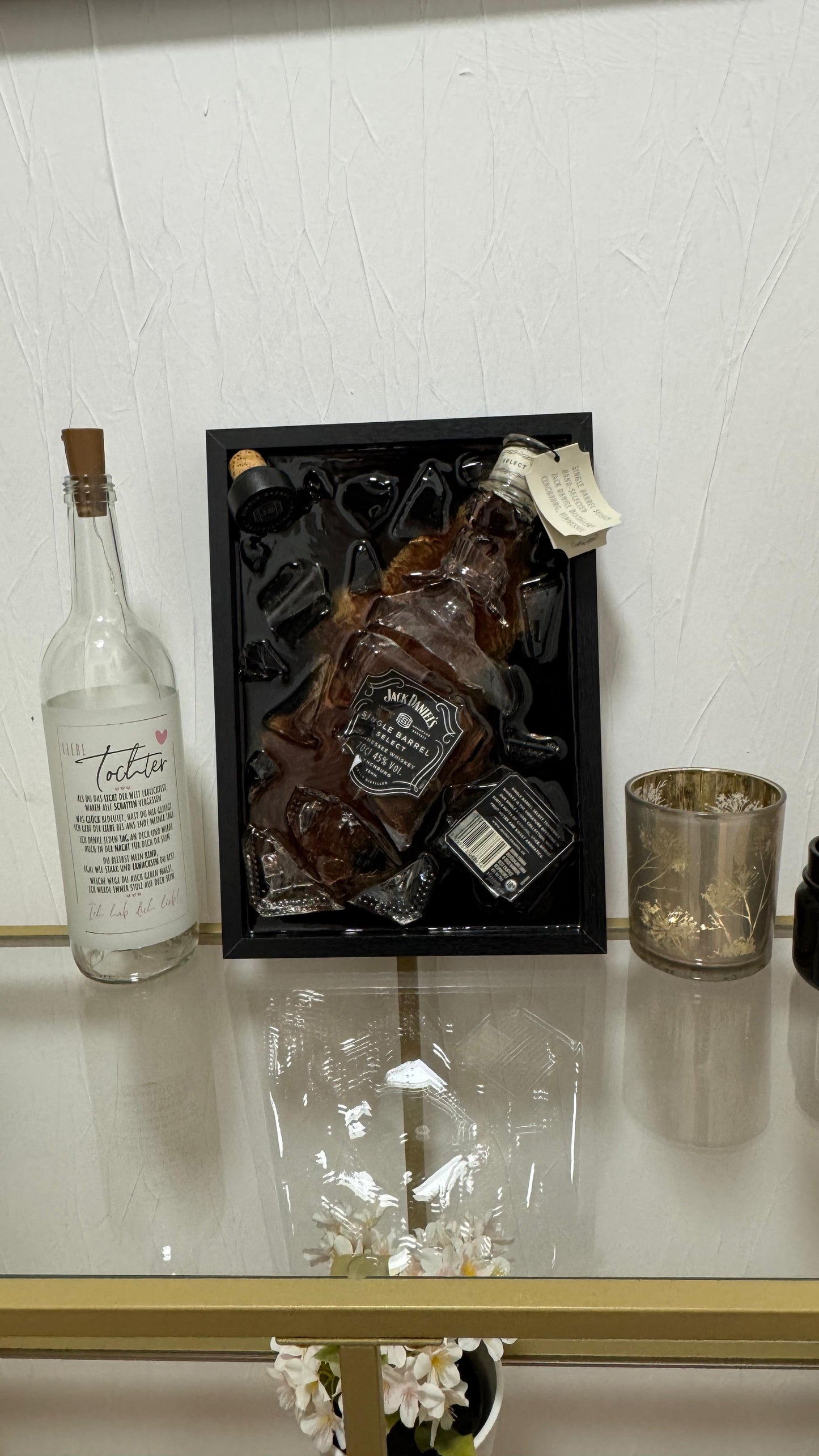 Eine Präsentation mit einer Jack Daniel's Single Barrel Select Whiskyflasche in einem schwarzen Rahmen mit Eis. Links eine klare Flasche mit Text und einem Korken. Ein kleines goldfarbenes Glas und eine schwarze Flasche sind ebenfalls vorhanden.