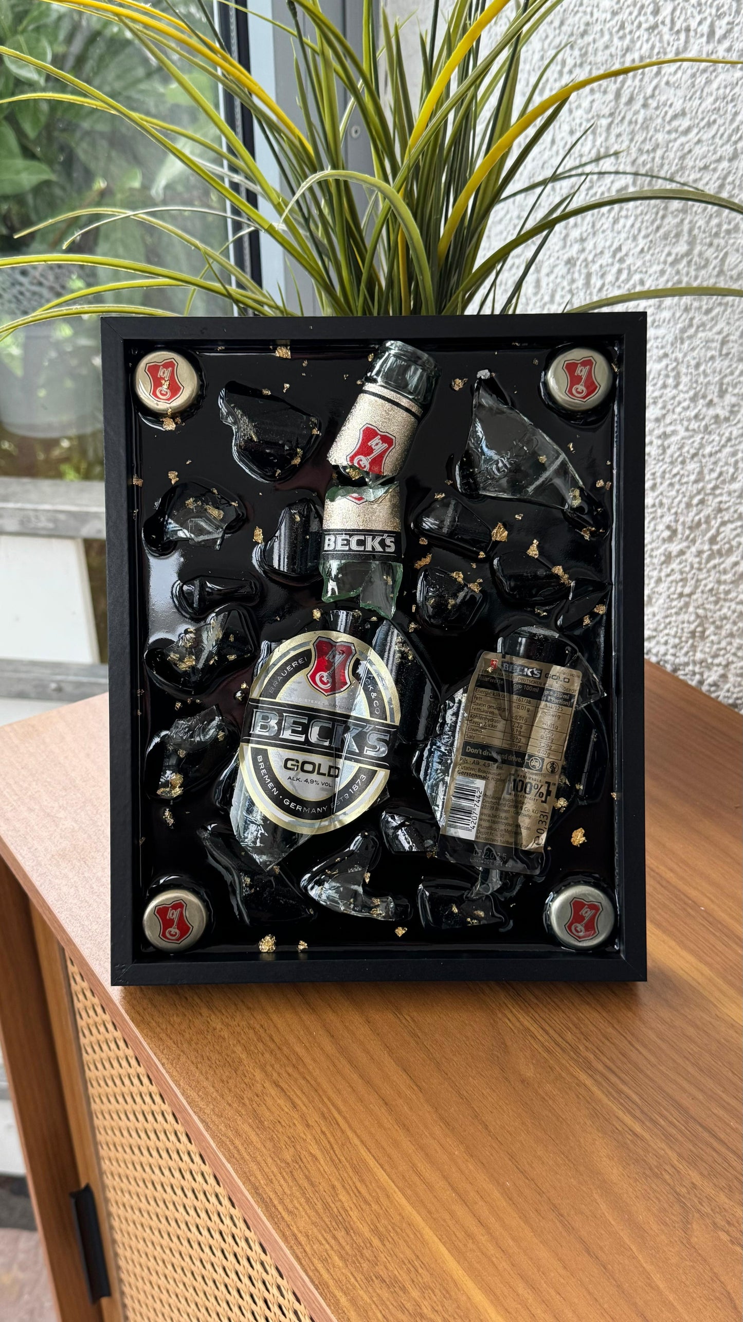 Handgefertigtes Wandbild mit zerbrochener Glasflasche, Kronkorken und goldfarbenen Details, in schwarzem Rahmen gegossen – kreatives Upcycling-Dekoobjekt.