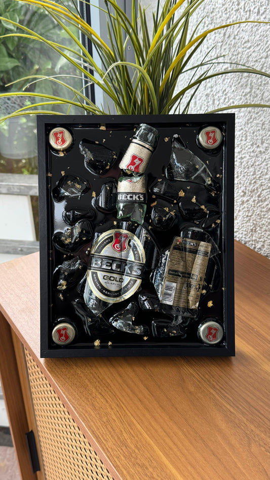 Handgefertigtes Wandbild mit zerbrochener Glasflasche, Kronkorken und goldfarbenen Details, in schwarzem Rahmen gegossen – kreatives Upcycling-Dekoobjekt.
