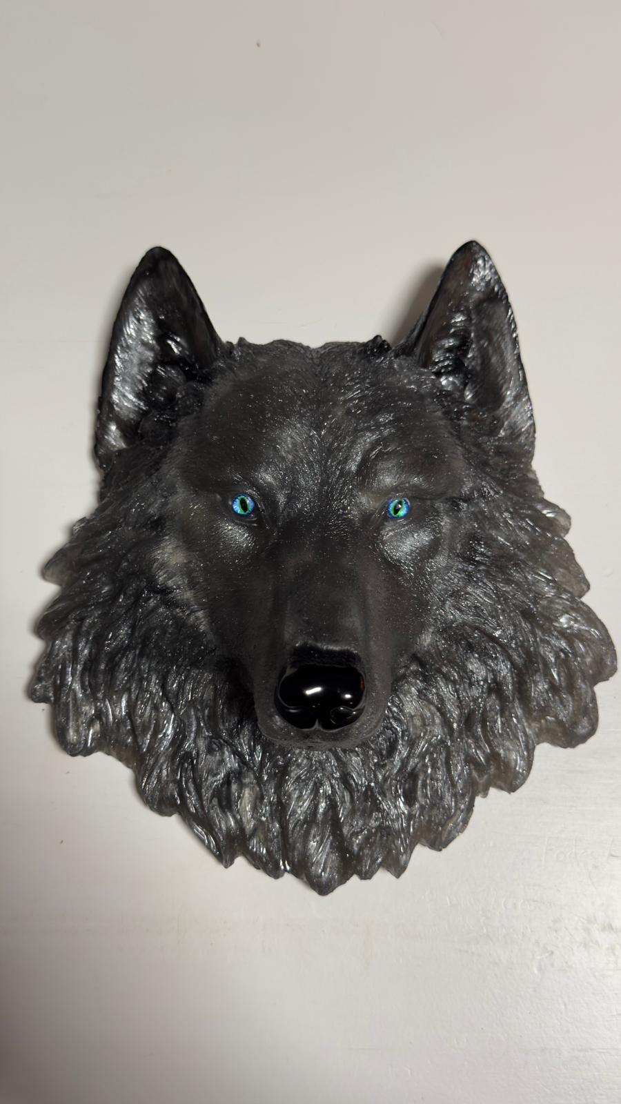 Könnte beinhalten: Detaillierte, dunkelgraue Wolfskopfskulptur mit auffälligen blauen Augen und einer glänzenden schwarzen Nase. Die Skulptur hat ein strukturiertes, fellartiges Aussehen und ist auf einer weißen Oberfläche montiert. Das Kunstwerk ist ein dekoratives Stück.