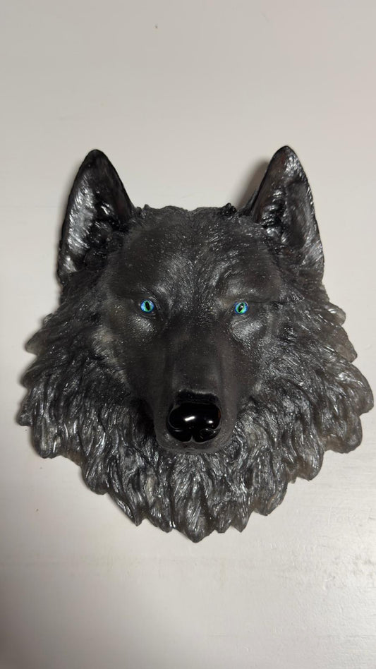 Könnte beinhalten: Detaillierte, dunkelgraue Wolfskopfskulptur mit auffälligen blauen Augen und einer glänzenden schwarzen Nase. Die Skulptur hat ein strukturiertes, fellartiges Aussehen und ist auf einer weißen Oberfläche montiert. Das Kunstwerk ist ein dekoratives Stück.