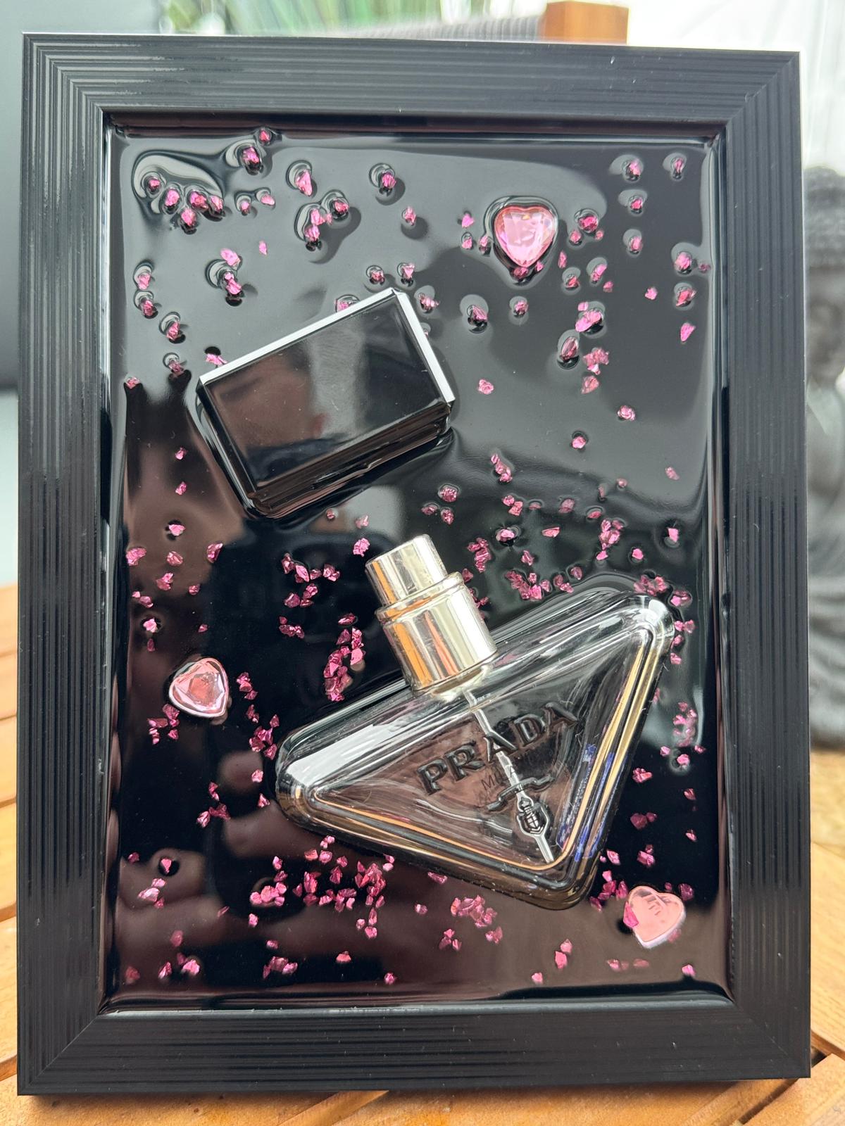 Ein gerahmtes Kunstwerk mit einem transparenten Parfümflakon mit dem Wort "PRADA", einer schwarzen Kappe und rosa herzförmigen Akzenten auf einem glänzenden schwarzen Hintergrund. Der Rahmen ist schwarz.